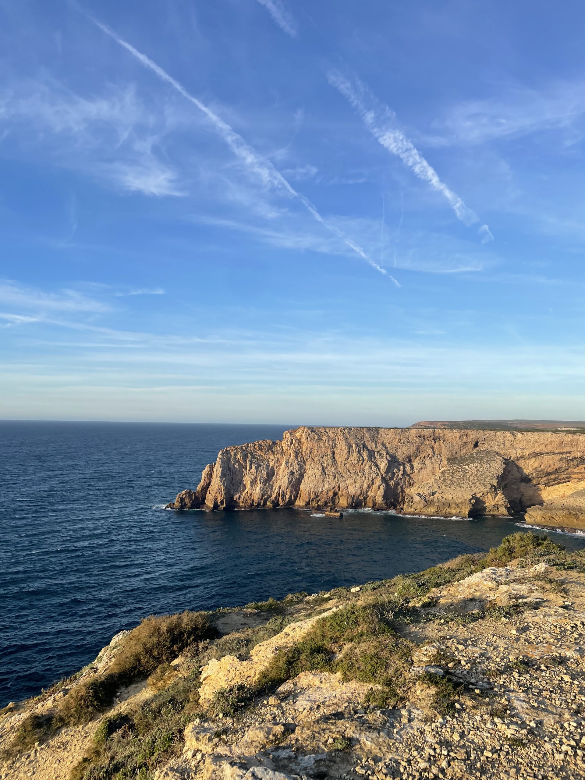 Impresionante acantilado rocoso bañando por el océano bajo un cielo azul con estelas de aviones, capturado al atardecer.