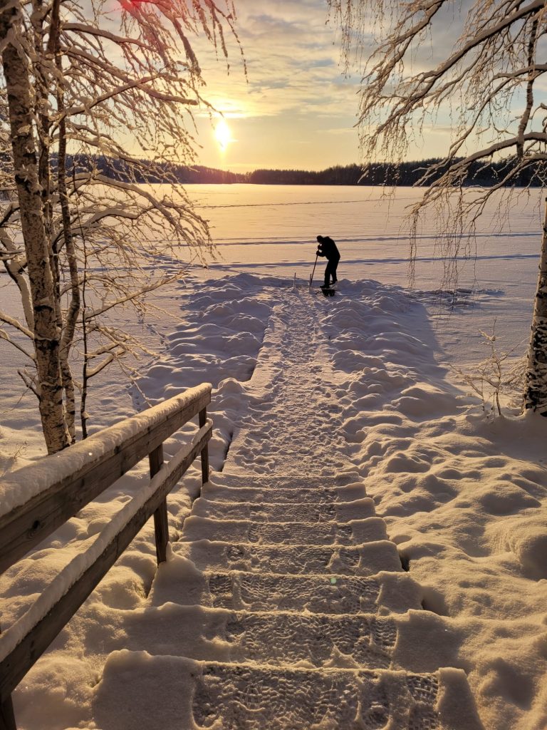 Escalera cubierta de nieve conduce a un lago congelado al atardecer, con una persona practicando esquí nórdico en el hielo